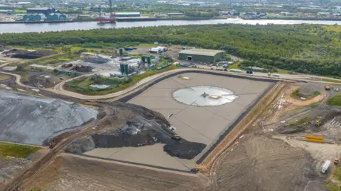 An aerial view of the Port Clarence landfill site. The site is grey and gravelled with lighter coloured tracks for vehicles. In the background can be seen a thick bank of trees and a river.