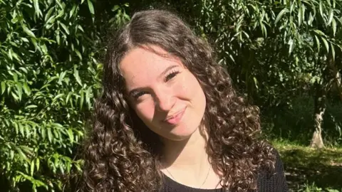 Lola Thompson A girl with dark curly long hair is pictured smiling and looking at the camera whilst standing in front of a tree.