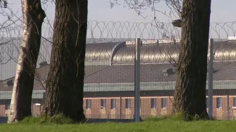 The prison surrounded by a security high fence and barbed wire and two trees in the foreground