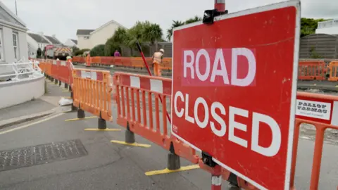 BBC A road closed red sign in front of rows of orange barriers on a road which is closed off.