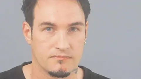 In a police custody photo, Ben Lewis has black hair, a black goatee beard, blue eyes and a small circular earring in one ear.