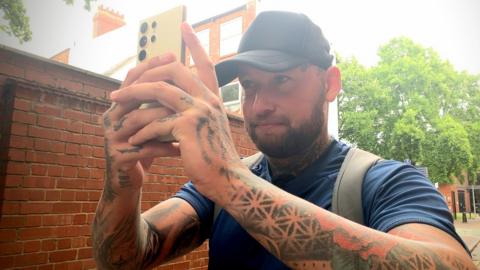 John Levesley holding up his phone taking a photo. He is wearing a black cap and blue t-shirt and has tattoos on his neck and arms.