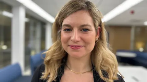 BBC/Helen Richardson Barbora Gray has long blonde wavy hair. She is smiling and wears a gold necklace.