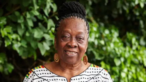 ACCI A woman is smiling at the camera, she has her hair braided in a bun and is wearing ornate circular earrings.