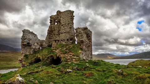 Ardvreck Castle is a ruin. There are three tall, stone-built walls and two cave-like openings below them. The castle is on a grassy knoll with Loch Assynt and low hills in the background. Grey clouds fill the sky above.