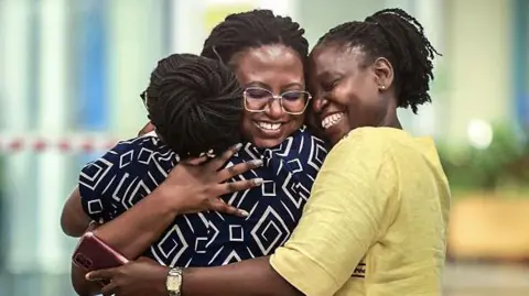 A person wearing a patterned dress is embracing two other people in an airport arrivals area, standing close together near a piece of luggage with a bag placed on top of it. The background shows a brightly lit terminal with glass walls and blurred interior details.