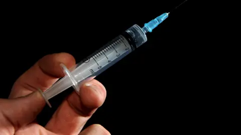 A close-up of a hand holding a clear plastic syringe at a 45 degree angle, pointing upwards to the right. The background is plain black.