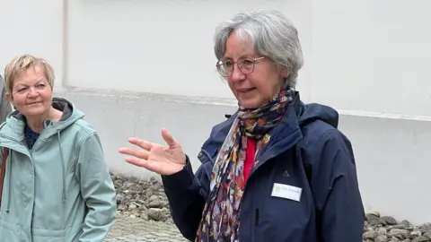 A group of people standing outside near a building with large windows. Gisela is wearing a dark jacket and a colorful scarf, appears to be speaking or gesturing. The others are dressed in casual outdoor clothing, including jackets and coats.