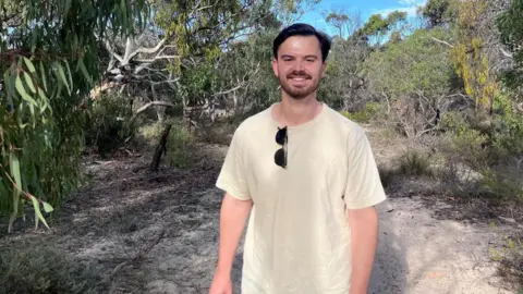 Handout Adam Laver, who has short, dark hair and dark facial hair. He is stood in a forested area of woodland.