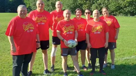 Supplied A group of men and women huddle together in matching red sports tops. The man at the front of the group is holding a rugby ball.