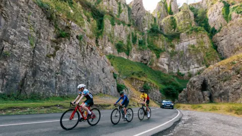 Bristol Bears Foundation Three people on bicycles and wearing cycling gear going down the road that runs through Cheddar Gorge. They are cycling towards the camera and there is a black car behind them in the distance. The gorge is towering above them making them look really small.