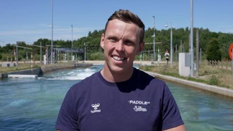 A smiling Joe Clarke wearing a blue T-Shirt looks directly towards the camera, squinting slightly from the sunshine, and standing in front of a slalom course at Lee Valley Water Centre
