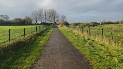 Daniel Mumby The Strawberry Line footpath near Ridge Road in Shepton Mallet. It is a wide tarmacked path with a metal fence lining either side, behind which are green fields and trees.