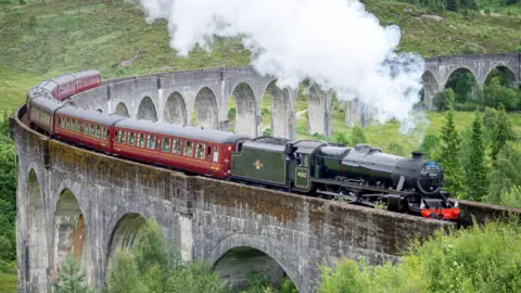 Steam rises from the train's black locomotive as it crosses the Glenfinnan Viaduct. The locomotive is hauling a line of carriages.