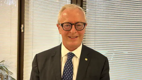 Mike Powell is wearing a suit and blue tie and has short white hair and black glasses.
