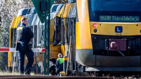 Reuters A Danish police officer in black uniform stands with his arms crossed next to the scene of a train collision. To his right, is a yellow train marked, a screen marking its direction reading 960R Tisvildeleje. Another man in a yellow hi-vi vest, can be seen standing on the tracks, next to the train