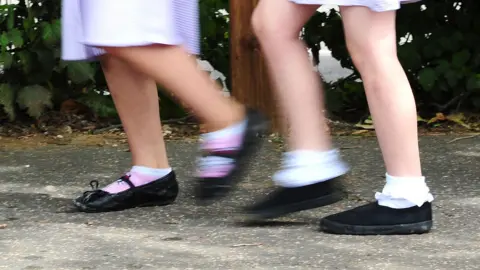 PA Media Two sets of children's legs can be seen mid-walk on a pavement. They are wearing short socks with black school shoes. The bottom of gingham school uniformed skirts can be seen just above the knees.