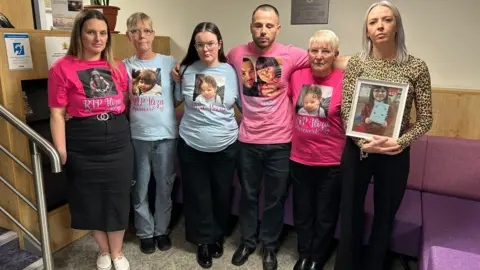 Julia Bryson/BBC A group of six people, with their arms around each other, stand in the entrance to a court and one is holding a framed photo of a young girl. Five of them wear t-shirts with her face and name on. 