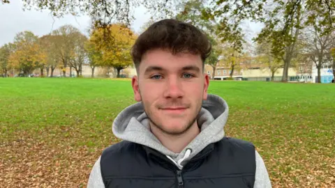 Sam Read/BBC A man in a grey hoodie and black gilet stands in a park with lots of trees in the background.