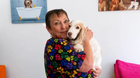 Marion Preston, a woman in a black top with multi-coloured dog paws printed on it. Holding a white and brown puppy.