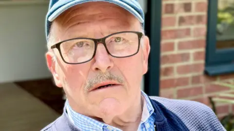 Close up photo of Ray sitting in the garden. He's wearing a blue baseball cap, black glasses and a blue shirt. He's looking into the camera with little expression on his face.