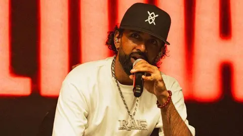 Manni Singh/@flyingbeardphotography Raf-Saperra, a British Asian man, wearing a white tshirt and black cap, with a silver chain which reads "RAF" around his neck. He is singing into a black microphone. Behind him on stage is a red LED light with block letters out of focus.