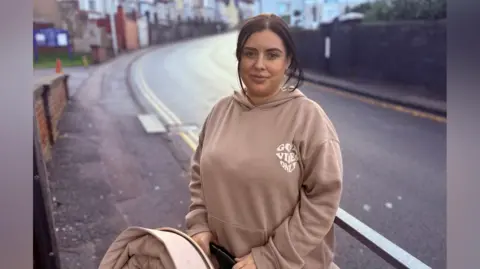 Demi Worboys pictured outside her temporary accommodation in Chatham, Kent. Demi is wearing a beige hoodie and is pushing a beige pram. The road in the background is blurred.