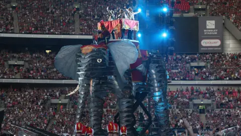Getty Images Take That stand together on a platform which sits on the back of a large mechanical elephant. The band members are waving and spreading their arms. A huge crowd is looking on in the background. 