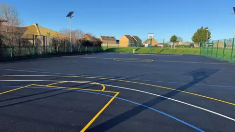 North Northamptonshire Council A multi-use games area, with markings on the ground. Houses are in the distance, as well as trees and bushes. There is high fencing all around the pitch. 