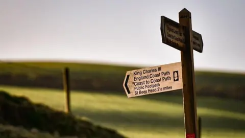 A wooden signpost, with one sign pointing to the King Charles III England Coast Path, and St Bees Head.
