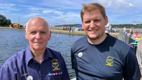 A man with grey hair stands next to a younger man in front of a body of water. The both wear Sara-branded shirts. They are looking at the camera and smiling.