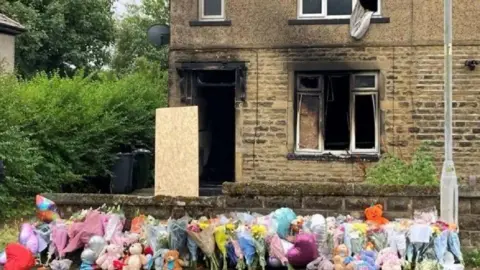 A large floral tribute rests against a garden wall in front of a house with visible fire damage.