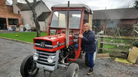 Dexta's On The Run Lesley is climbing up into her red tractor, which has small old fashioned wheels at the front. Her tractor has a cab to sit in.