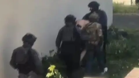 Blurred still from footage showing the backs of unidentifiable counter-terror officers and special forces arresting a man whose face cannot be seen. He is bent downwards walking along with the officers