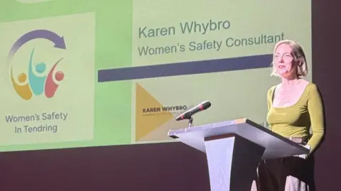 Tendring District Council A woman standing at a podium. She is wearing a green top and talking into a microphone.
She has short blonde hair that comes just below her ear and is tucked behind her ear. Behind her is a slide that reads "Karen Whybro Women's Safety Consultant"