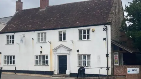 A photo of the front of Essex House on Fore Street building which has structural damage. The building is white with seven windows and a black door. 