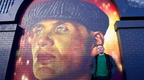 A man wearing a green jumper and a blue coat. He is standing in front of the mural of Thomas Shelby which has an orange and red painted background. Thomas is wearing a black flat-cap and staring into the distance in the mural.