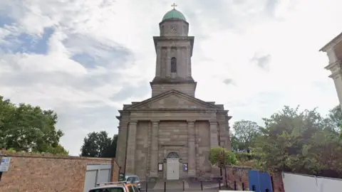 A large stone church with a large tower with a blue dome on the top. It is at the end of a road with vehicles parked either side, houses on the right and a wall on the left. Below the dome there is a clock face