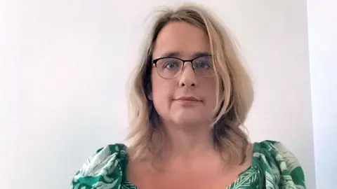 Emma Harris Emma Harris wearing glasses and a green floral top against a white wall