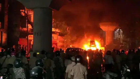 A street on fire, seen with people, including police, overlooking the blaze. 