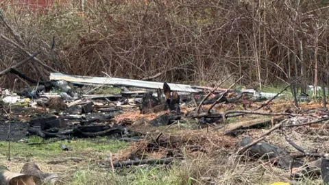 Mike Sib A pile of half burnt metal and wood. There is blackened ash on the ground near the pile of waste.