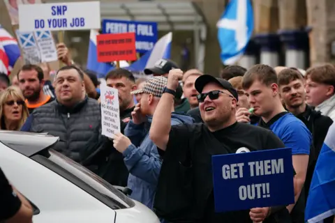 PA Media A group of people, mostly men, protesting against asylum seekers being housed in hotels. Many are holding signs - one man in the foreground has a sign that reads "get them out!!"