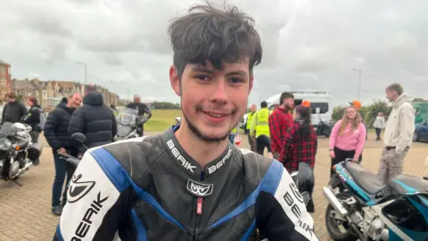 William wearing black and blue motorcycle leathers with short curly hair, smiling at the camera. There are people loitering in the background in a car park with a few other motorbikes in view.