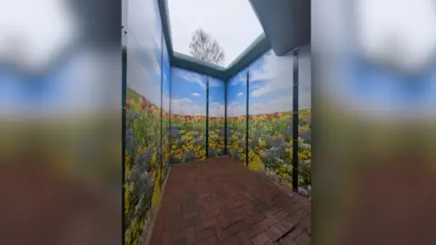 Avon and Wiltshire Mental Health Partnership NHS Trust An enclosed room with flowers and grass with brick flooring. It is a peaceful scene.