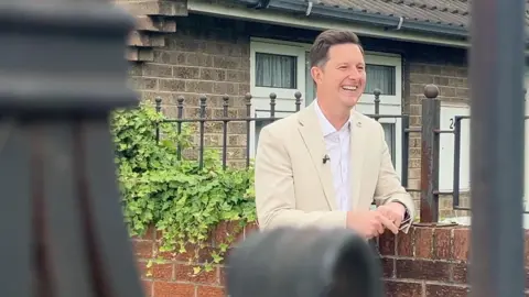BBC/James Vincent Doncaster East and Isle of Axholme MP Lee Pitcher smiles as he leans on a low red brick wall. He is wearing a cream suit jacket with a white shirt. 