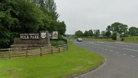 Google Maps A Google Maps screenshot of the Ulster American Folk Park sign, which is situated beside a main road. There is a car driving towards the sign, with a number of fields on the right hand side of the image and large trees planted around the sign.