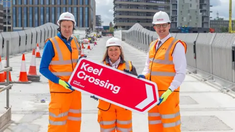 Councillor Kevin Johnston, councillor Lindsey Leonard and councillor Michael Mordey, hold a red sign reading Keel Crossing at the new footbridge. They are wearing orange protective clothing and white helmets. The grey concrete structure is still under construction. There are more new building on its other side, including the City Hall. Keel Square is also visible in the distance.