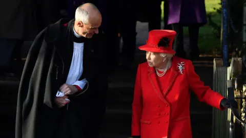 BEN STANSALL/AFP via Getty Images The late Queen is on the right. She is wearing a red dress, a red coat and a red hat. She is holding a railing on the right and speaking to a reverend, who is on the left. He is leaning towards her. 