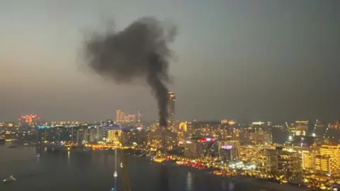 Night scene of the Dubai shoreline showing a plume of thick black smoke rising from a group of buildings. 