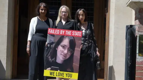 Jamie Niblock/BBC Victoria Sebastian stands outside the front of Arundel House in London holding a big poster of her daughter, with the words "Failed by the state" and "We love you". She has two solicitors beside her. They are all wearing black.